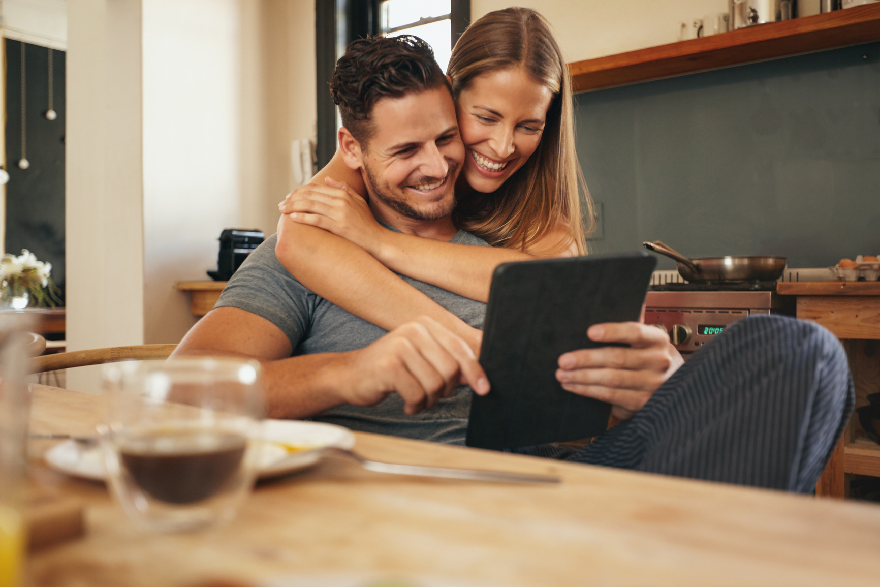 Couple smiling as they read a tablet computer together in morning in the kitchen. Young man and woman catching up on social media smiling. Woman hugging her boyfriend holding tablet PC.